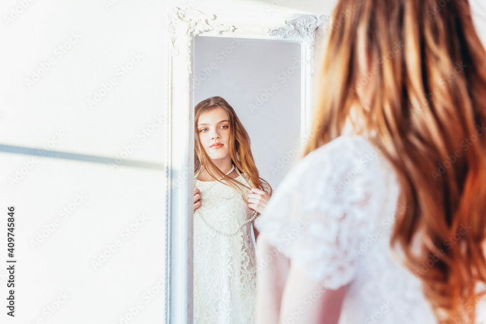Beauty makeup morning rutine love yourself concept. Young teenage girl looking at reflection in mirror. Young positive woman wearing white dress posing in bright light room against white wall Beauty makeup morning rutine love yourself concept. Young teenage girl looking at reflection in mirror. Young positive woman wearing white dress posing in bright light room against white wall