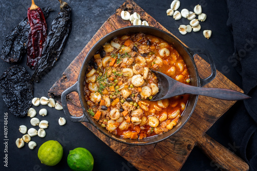 Traditional slow cooked Mexican pozole rojo soup with ground minced beef, hominy maiz and dry ancho paprika offered as top view in cast-iron roasting dish on old rustic wooden board photo