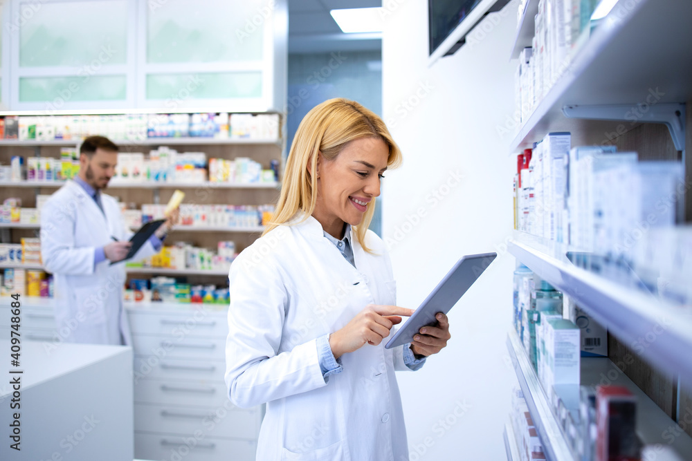 Beautiful blonde pharmacist typing on tablet computer and selling medicine online in drugstore.