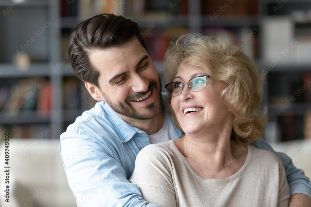 Head shot close up happy young caucasian man embracing from back sincere smiling senior old retired mother, showing love care gratitude at home, different generations family relations concept. Head shot close up happy young caucasian man embracing from back sincere smiling senior old retired mother, showing love care gratitude at home, different generations family relations concept.
