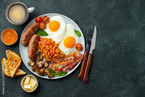 Full English breakfast on a plate with fried eggs, sausages, bacon, beans, toasts and coffee on dark stone background. With copy space. Top view