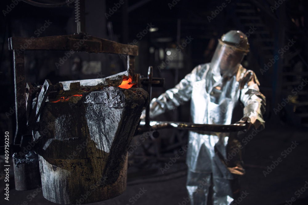 Foundry worker in aluminized protective fire suit pushing bucket with molten liquid iron in smeltery.