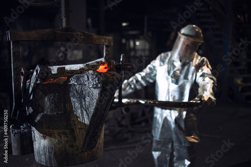 Foundry worker in aluminized protective fire suit pushing bucket with molten liquid iron in smeltery. © littlewolf1989