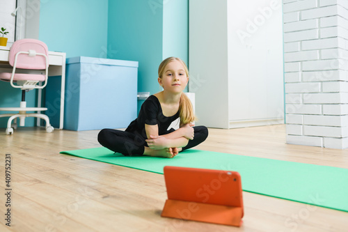 Elementary school girl put sport mat and tablet on floor, wearing black leotard before start online remote gymnastic class during self-isolation at home