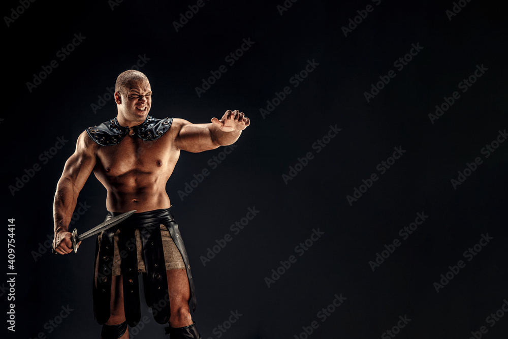 Severe barbarian in leather costume with sword. Portrait of balded muscular gladiator. Studio shot. Black background.