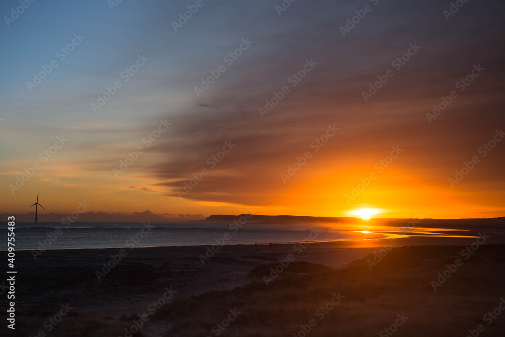 Calm ocean with powerful sunrise at deserted sandy beach with reflections. Wind turbine in background