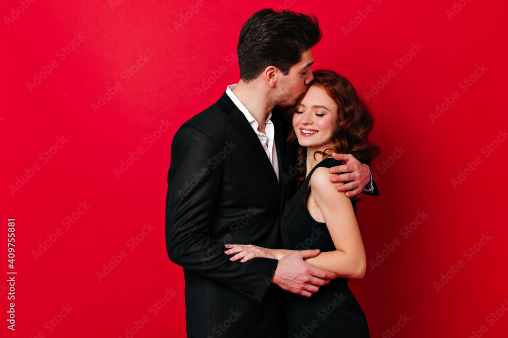 Brunette man kissing wife in forehead. Studio shot of loving couple isolated on red background