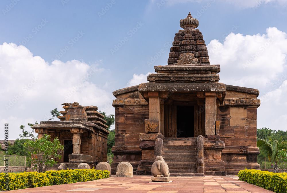 Aihole, Karnataka, India - November 7, 2013: Huchchimalli Gudi or Temple. Brown stone main building with small adjacent shrine under blue cloudscape and green foliage in back. Nandi statue.