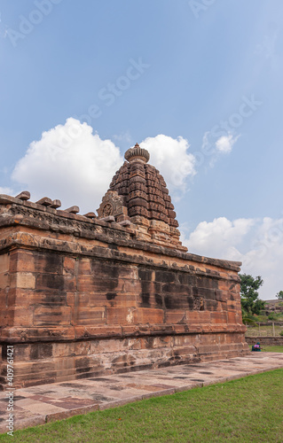 Aihole  Karnataka  India - November 7  2013  Huchchimalli Gudi or Temple. Side red stone wall with sculpted vimanam towering over it under blue cloudscape. Green grass in front.