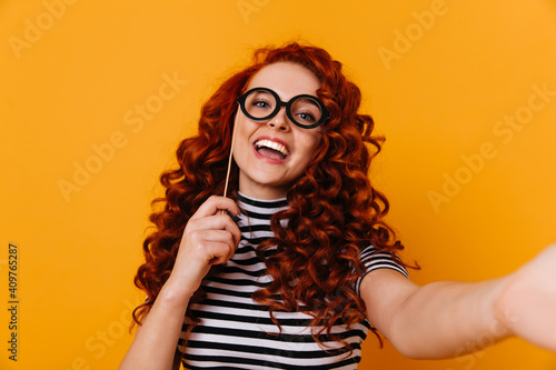Naughty young woman in striped T-shirt poses with toy glasses and takes selfie in orange studio © Look!