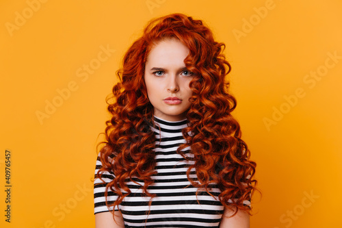 Blue-eyed curly girl with sad face expression looks into camera. Photo of redhead woman in striped top on orange background