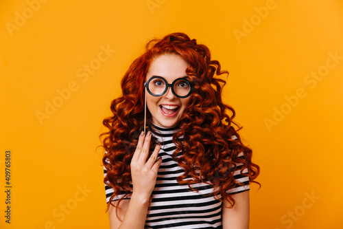 Charming woman with blue eyes and red hair is holding glasses model and is smiling on orange background