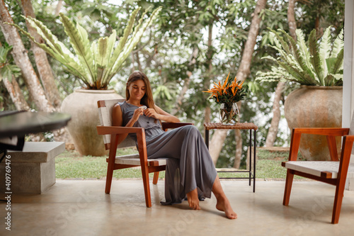 Home lifestyle woman relaxing on outdoor patio living room, smell the flower, sit on rocket chair. Young woman sitting on chair in green garden terrace