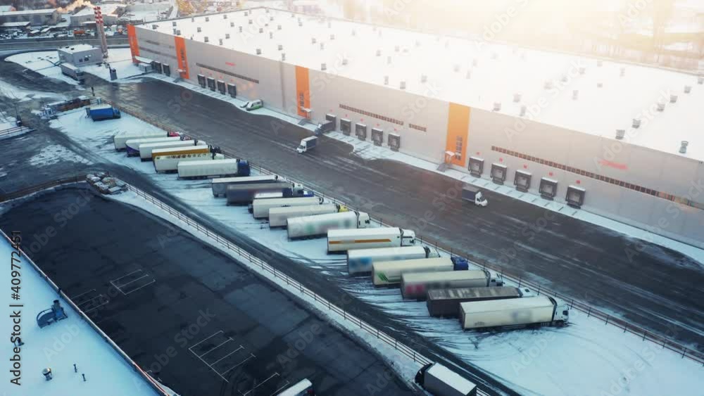 Semi-trailers trucks stand on a parking lot and wait for load and unload goods at the warehouse ramps in the logistics park. Aerial view at winter sunset Semi-trailers trucks stand on a parking lot and wait for load and unload goods at the warehouse ramps in the logistics park. Aerial view at winter sunset