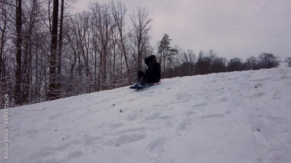 A woman wearing black winter clothes, boots and gloves is on a snow sled, accerelating downhill on snow covered slope into the woods. Panning footage with winter landscape. A woman wearing black winter clothes, boots and gloves is on a snow sled, accerelating downhill on snow covered slope into the woods. Panning footage with winter landscape.