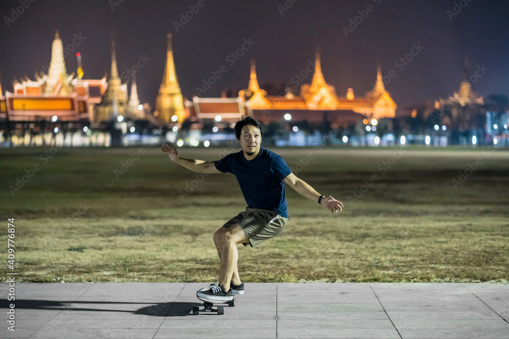 Asian Cheerful man playing surfskate or skate board in outdoor Park at night over photo blur of Bangkok Grand Palace,Thailand, low light, extream sport, healthy and exercise,fashion in covid19 concept