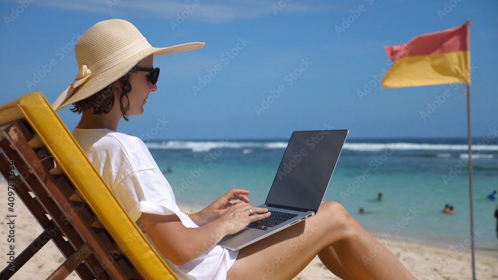 Side view of a woman in a straw hat sitting in a deck chair on a beach types on her laptop 