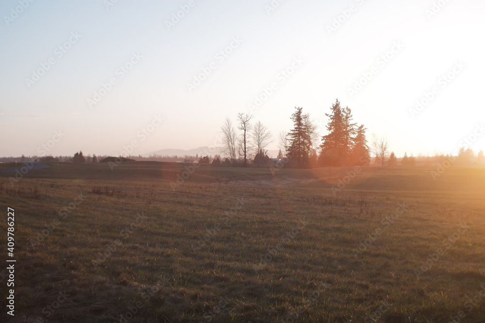 Sunset over a wide field in winter bathed in warm light with bare trees and evergreens in front of hills in the distance Sunset over a wide field in winter bathed in warm light with bare trees and evergreens in front of hills in the distance