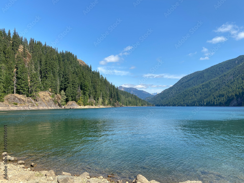 Mountain lake surrounded by forested slopes and a rocky beach on a sunny summer day, with a snow capped peak visible in the far distance