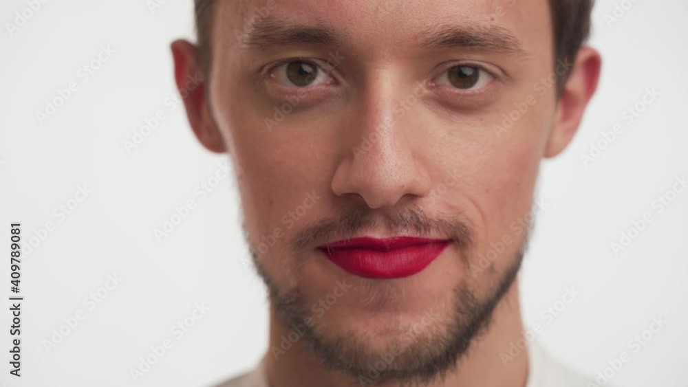 Beautiful young man with dark beard, brown eyes, moustache wear makeup – red lipstick on lips, looks around, stare camera isolated on white wall background close-up. Portrait of one Caucasian sweet bo Beautiful young man with dark beard, brown eyes, moustache wear makeup – red lipstick on lips, looks around, stare camera isolated on white wall background close-up. Portrait of one Caucasian sweet bo