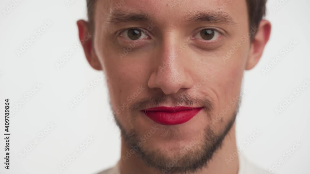 Portrait of glamorous man with beard, painted lips with bright red lipstick. Close-up of bearded guy smiles, winks with brown eyes, flirts, looks straight ahead like in a mirror on white background. Portrait of glamorous man with beard, painted lips with bright red lipstick. Close-up of bearded guy smiles, winks with brown eyes, flirts, looks straight ahead like in a mirror on white background.