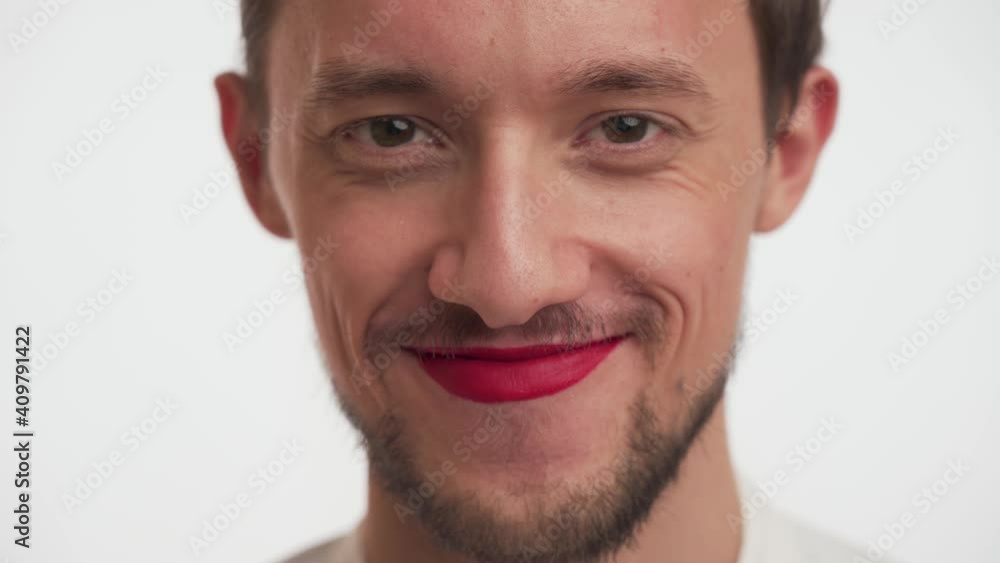 Close up face of serious confident young man with dark brown beard, moustache wear makeup – red lipstick on lips, looks at the camera, smiles, laughs sincerely isolated on white background. Happy guy Close up face of serious confident young man with dark brown beard, moustache wear makeup – red lipstick on lips, looks at the camera, smiles, laughs sincerely isolated on white background. Happy guy