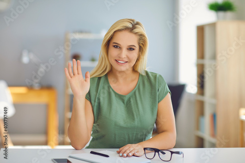 Beautiful positive young blond woman sitting and greeting somebody with hand during online meeting or video call at home or in office. Freelance, distant working, online communication concept