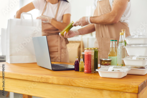 Healthy food delivery. Close up of containers and glass jars with healthy food on table near laptop on background of cafe workers. People in the background collect customer orders in packages.
