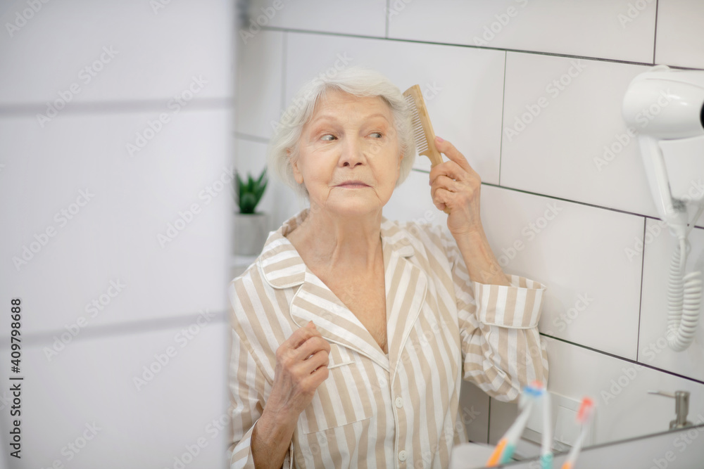 Gray-haired woman brushing her hair in the bathroom and feeling good Gray-haired woman brushing her hair in the bathroom and feeling good