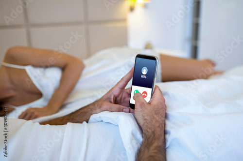 Cheating unfaithful man lying with mistress in hotel bed. Call from wife to mobile phone. Cheater having affair with secret lover and relationship with another woman. Infidelity and love triangle. photo