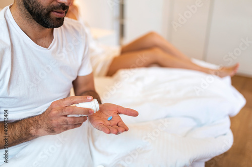 Man Suffering From Erectile Dysfunction Holding Pill In Hand And His Wife In Background. man takes viagra before sex, woman behind him in bed.