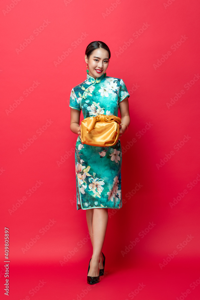 Happy smiling Asian woman in traditional costume giving gold gift box in red isolated studio background for Chinese new year concepts
