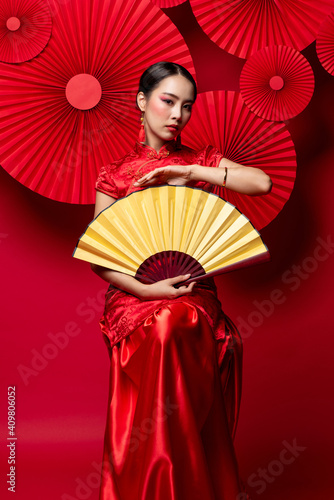 Beautiful glamourous Asian woman in traditional Chinese dress with colorful face make up holding golden fan in oriental style red background