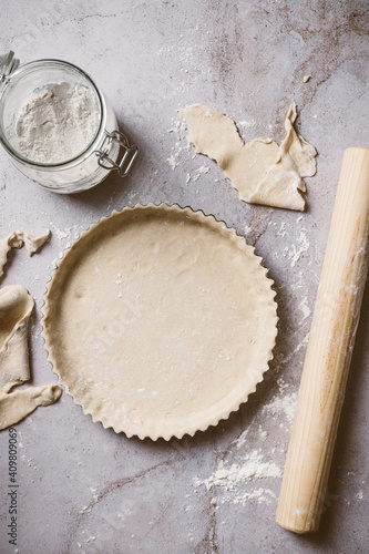 Overhead shot showing process of preparing a pie or fruit tart over a natural stone background. The photo includes flour, pieces of dough and a rolling pin.