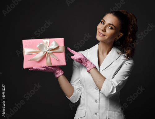 Young pretty brunette woman doctor nurse in latex gloves and uniform medical gown stands holding and pointing at gift box with ribbon over dark background. Gynecology concept photo
