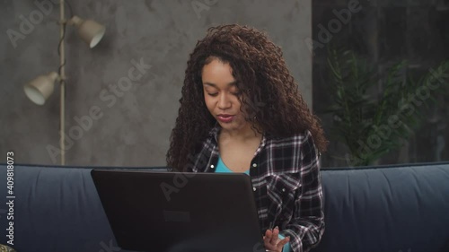 Portrait of attractive carefree curly african american woman sitting on couch in domestic room, chatting online using laptop pc, expressing positive emotions during exciting conversation in network. photo