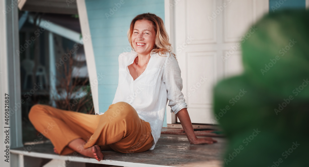 Beautiful happy adult blonde woman sitting on a terrace in a country house with a garden on a spring summer day