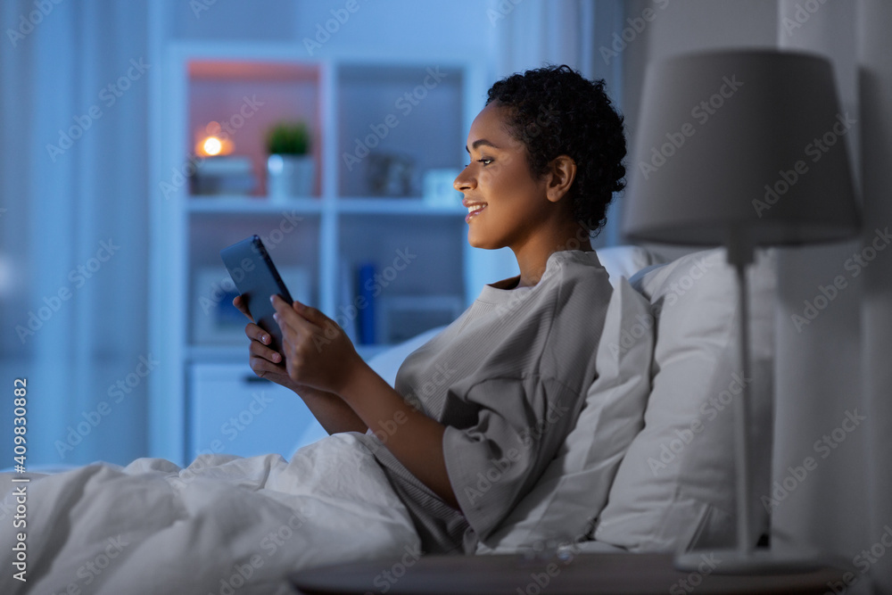 technology, internet and people concept - happy smiling young african american woman with tablet pc computer lying in bed at home at night technology, internet and people concept - happy smiling young african american woman with tablet pc computer lying in bed at home at night
