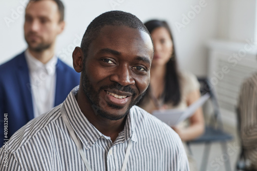 Head and shoulders portrait of African-American businessman looking at camera and smiling while sitting in audience at business conference or seminar, copy space