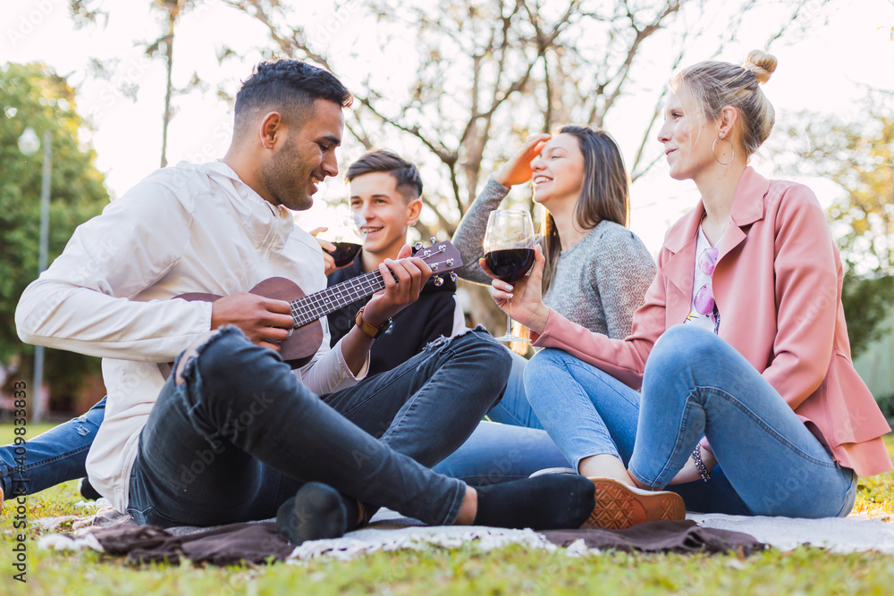 A group of friends sitting on the grass enjoying the outdoors - A young man playing the ukulele while his friends enjoy their music and a good wine.