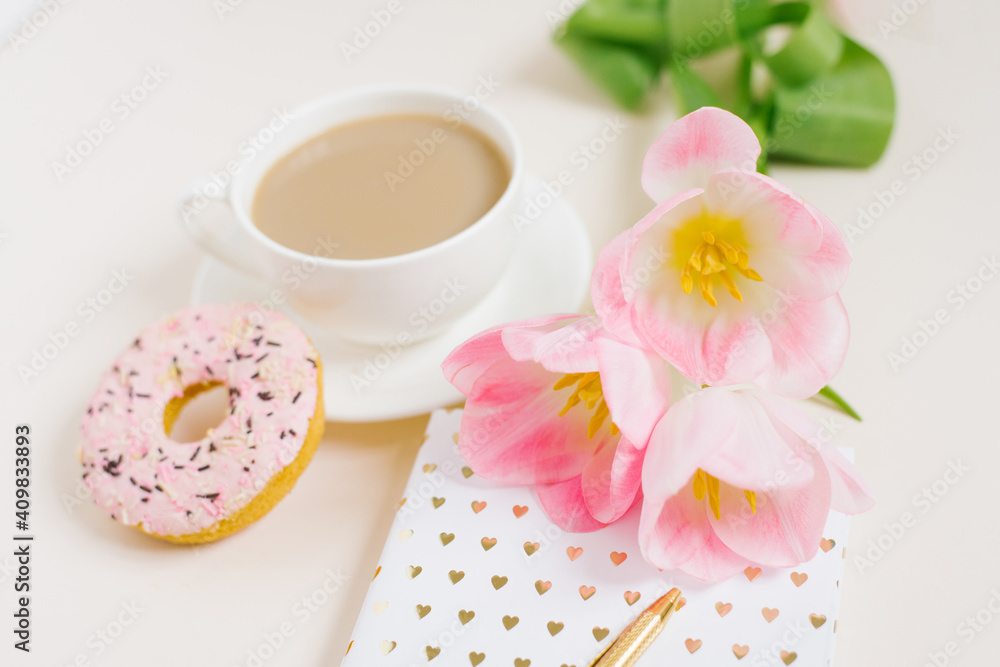 Spring breakfast for a blogger or office worker: a cup of coffee, a donat, a bouquet of pink tulips, a notebook with a pen Spring breakfast for a blogger or office worker: a cup of coffee, a donat, a bouquet of pink tulips, a notebook with a pen