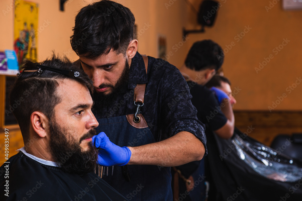 The concentrated barber who stands at his client's side and uses sterile instruments while cutting his client's hair - An expert barber who enjoys his work in the barber shop . The concentrated barber who stands at his client's side and uses sterile instruments while cutting his client's hair - An expert barber who enjoys his work in the barber shop .