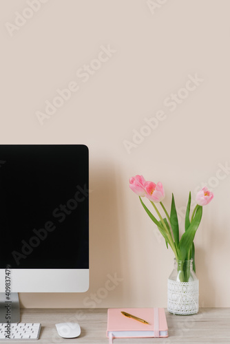 A computer monitor, a bouquet of pink tulips, and a notebook with a pen on a light wooden table against a beige wall. The workplace of a freelancer or student
