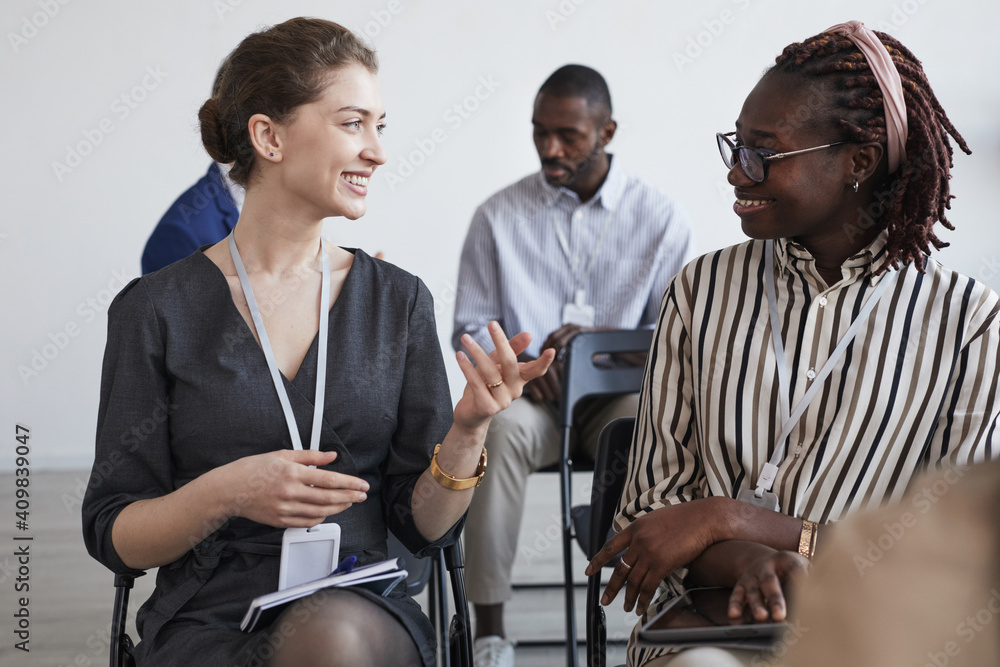 Obraz premium Portrait of two smiling businesswomen talking while sitting on chairs in audience at business conference or seminar