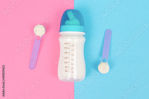 Bottle of milk for newborn baby and spoon with powdered infant formula on light pink blue table background