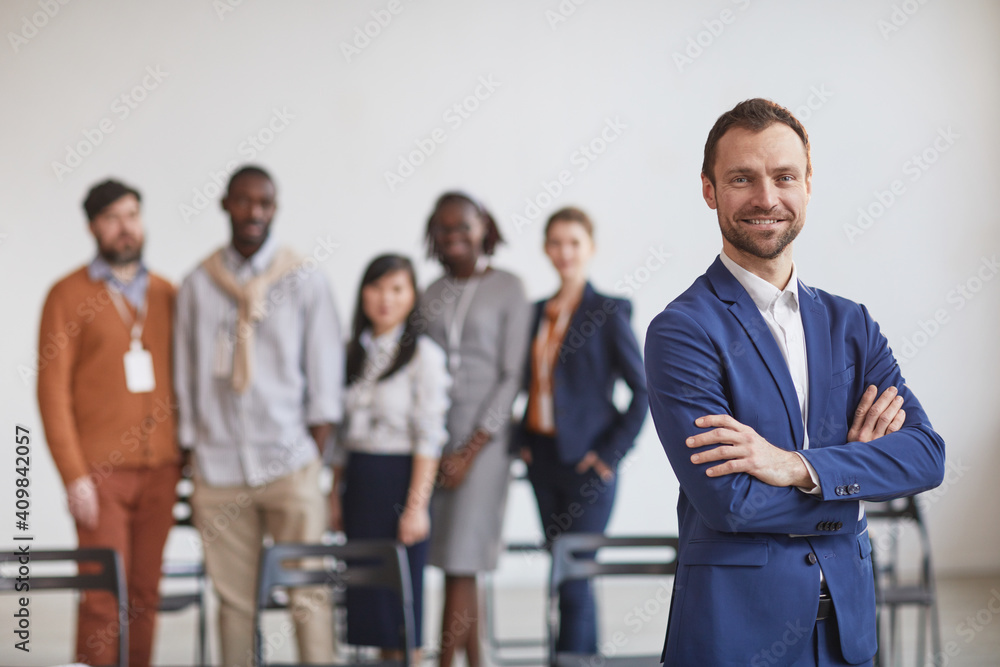 Waist up portrait of successful businessman standing with arms crossed against multi-ethnic team in background, copy space Waist up portrait of successful businessman standing with arms crossed against multi-ethnic team in background, copy space