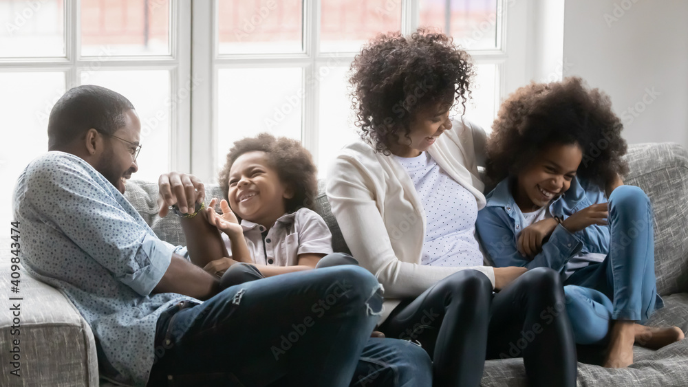 Close up overjoyed African American parents with kids tickling, having fun, sitting on cozy couch at home, happy mother and father with little son and daughter cuddling, involved in funny activity Close up overjoyed African American parents with kids tickling, having fun, sitting on cozy couch at home, happy mother and father with little son and daughter cuddling, involved in funny activity