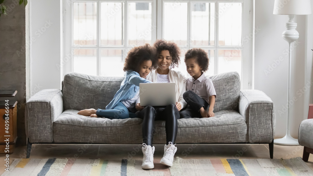 Happy African American woman with kids using laptop at home, smiling mother with little daughter and son looking at laptop screen, shopping or chatting online, watching video, sitting on couch Happy African American woman with kids using laptop at home, smiling mother with little daughter and son looking at laptop screen, shopping or chatting online, watching video, sitting on couch