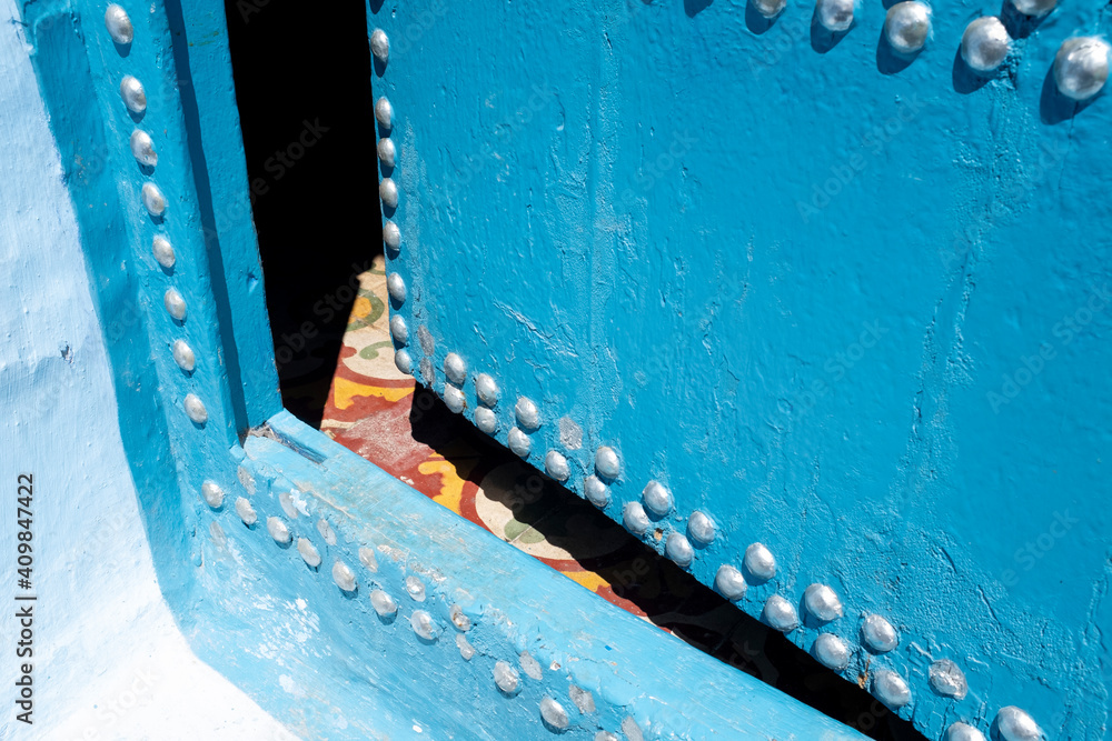Puerta vieja de madera azul con remaches metálicos entreabierta dejando ver azulejos de colores cálidos en el suelo. Chauen, Marruecos.