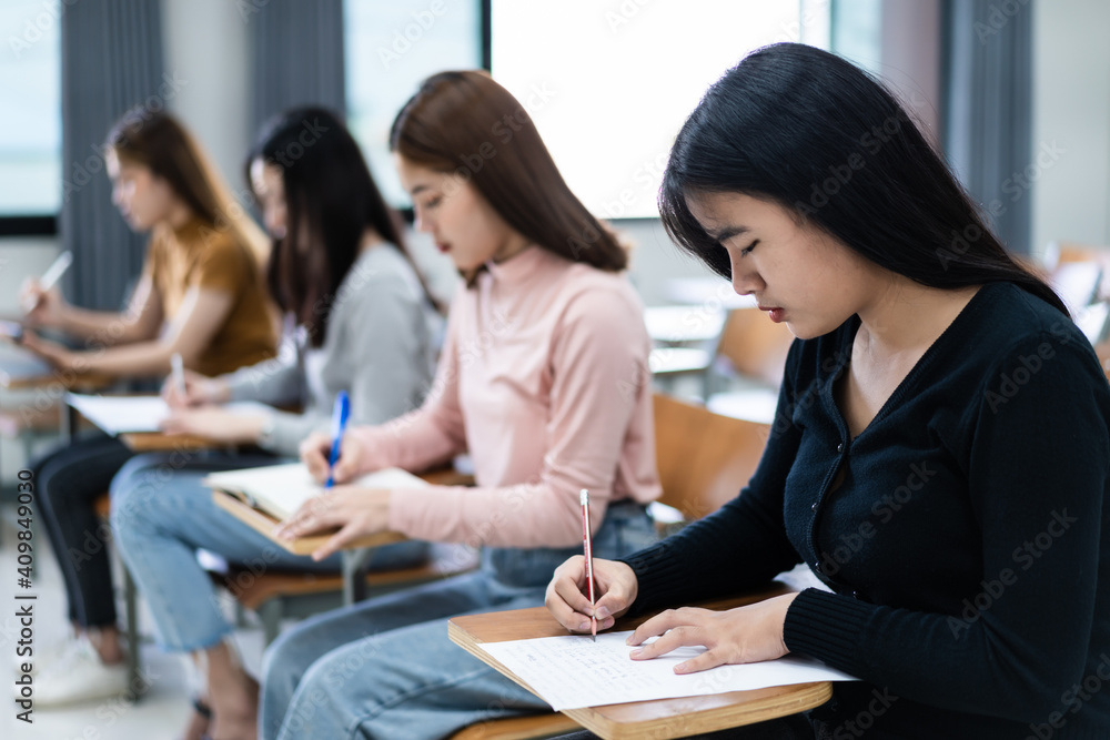 Selective focus of the teen college students sitting on lecture chair in classroom write on examination paper answer sheet in doing the final examination test. Female students in the student uniform. Selective focus of the teen college students sitting on lecture chair in classroom write on examination paper answer sheet in doing the final examination test. Female students in the student uniform.